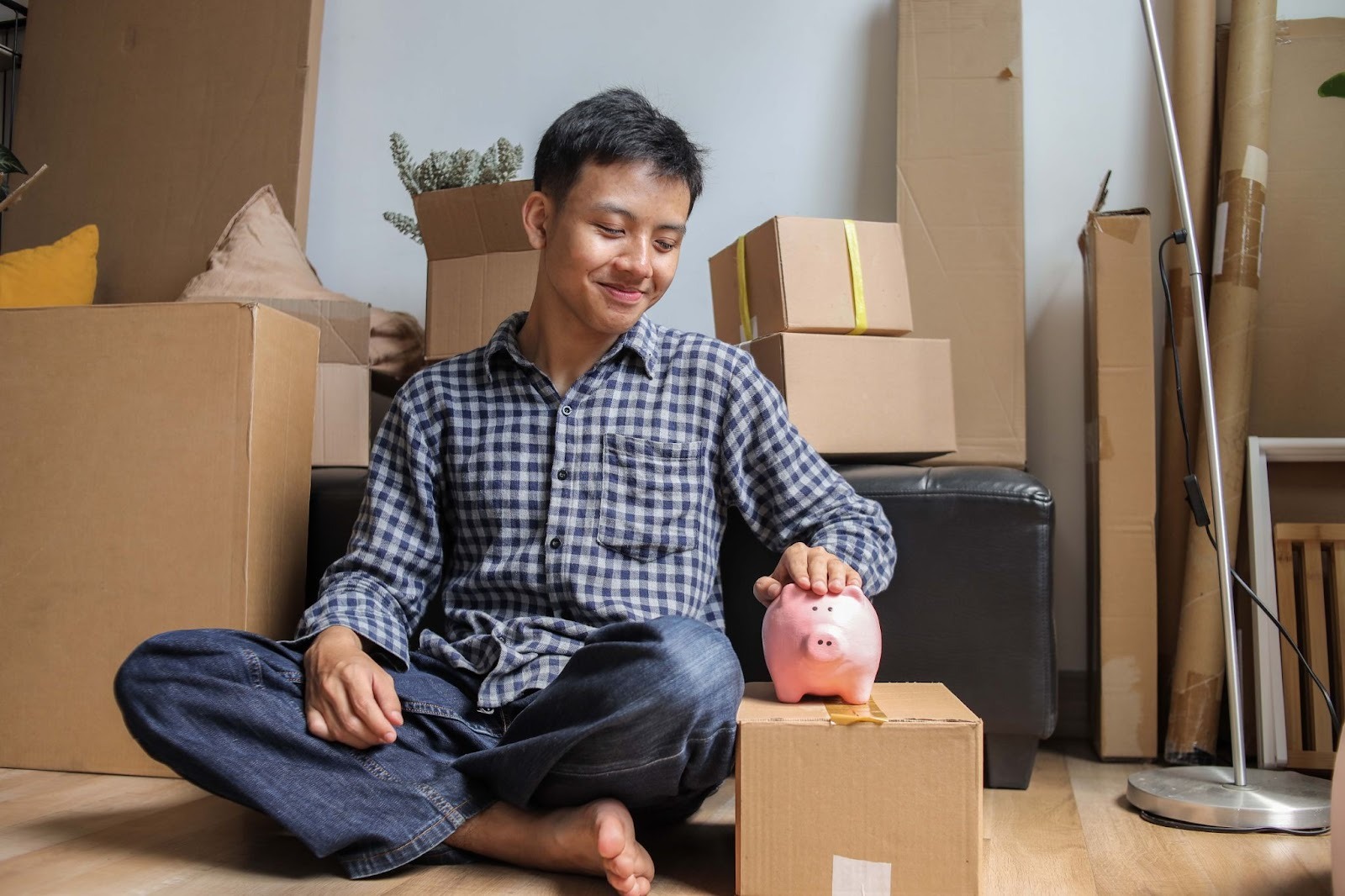 A young man sitting on the floor pats his piggy bank while leaning against boxes after relocation