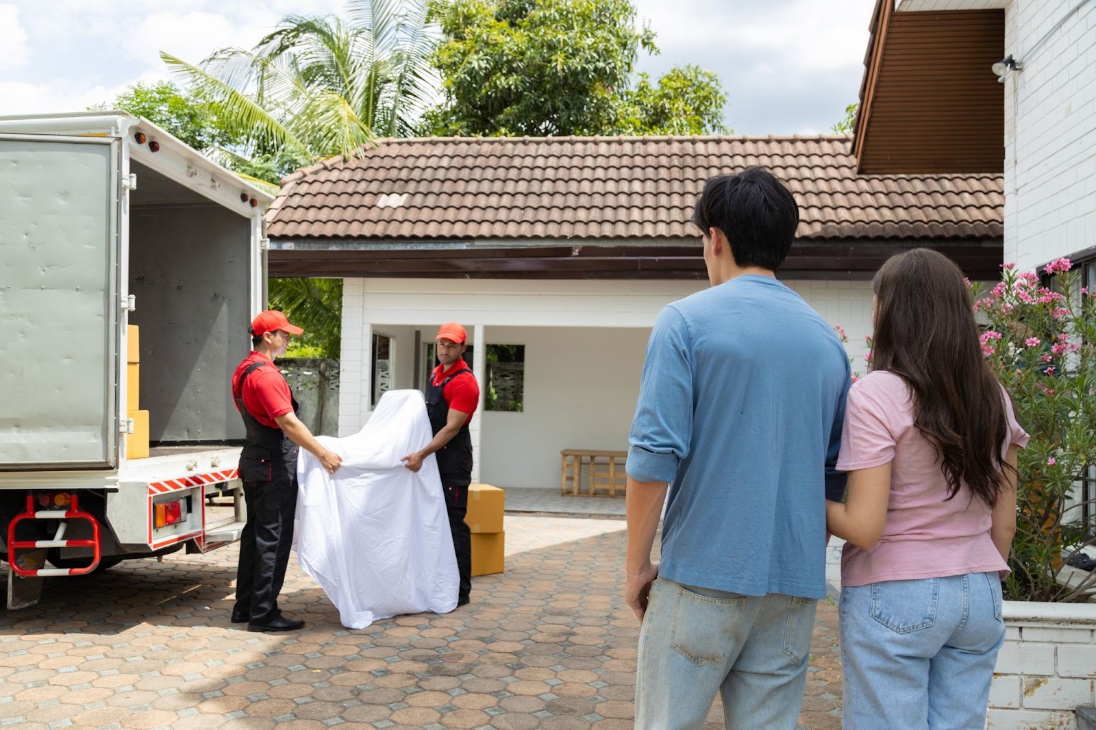 Residential movers unload boxes and furniture from a pickup truck while a young couple watches nearby