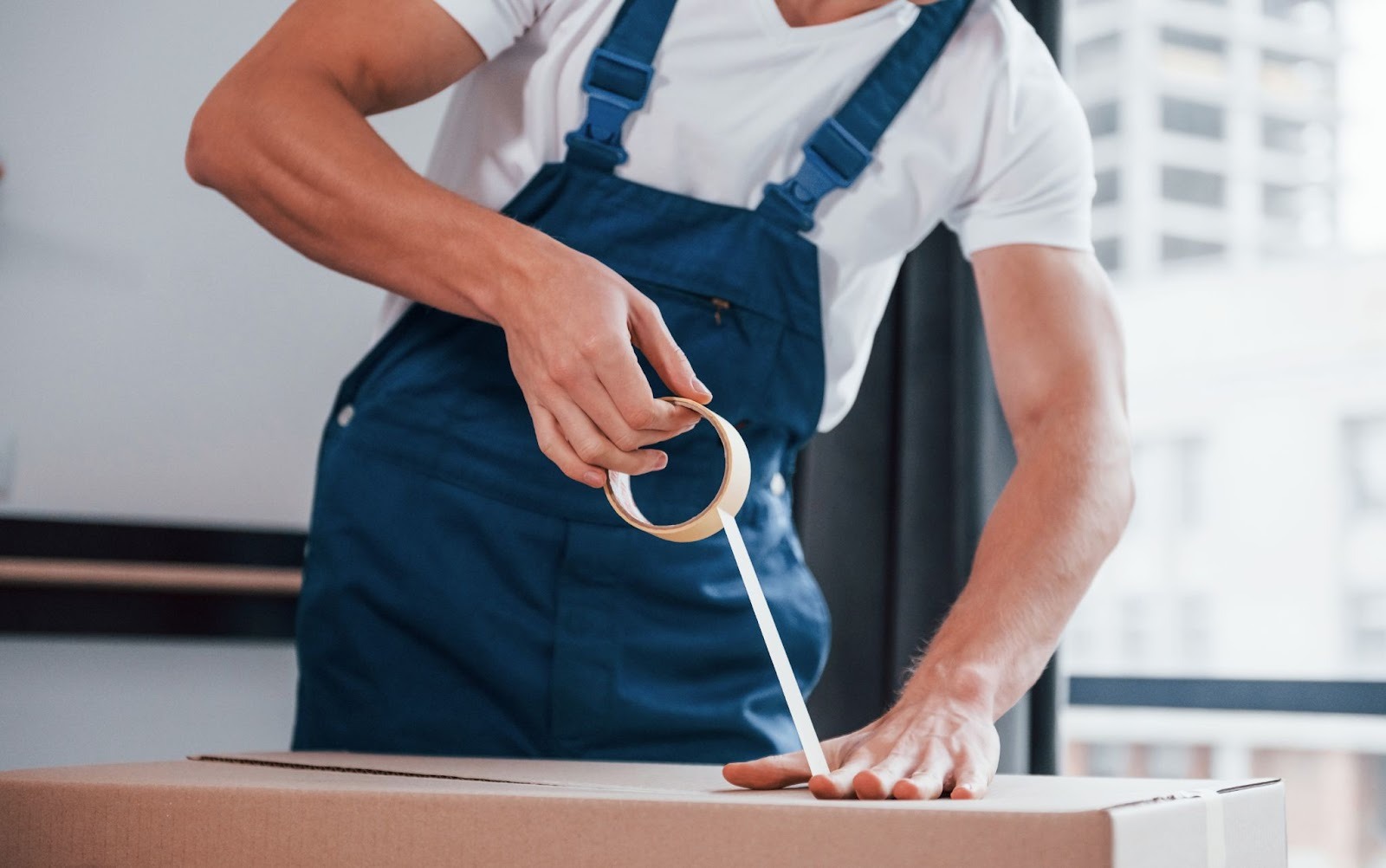 A professional mover in a blue uniform carefully sealing and labeling a moving box inside a room during relocation preparation
